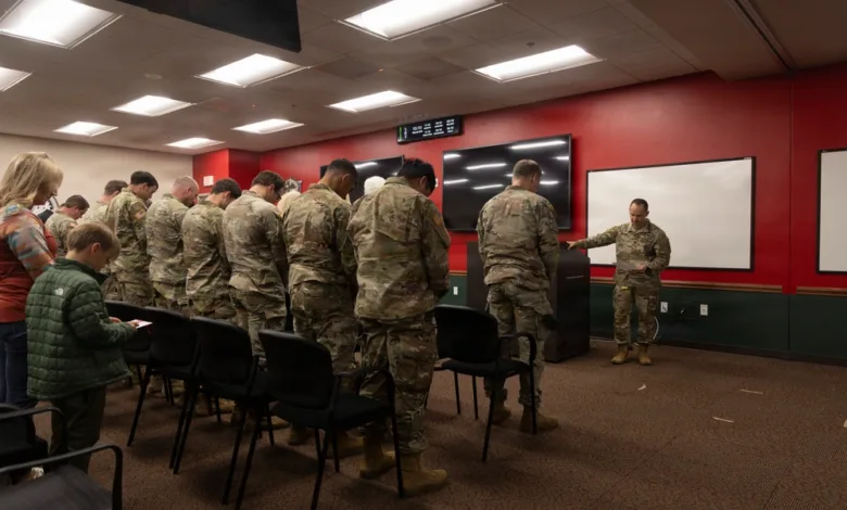 Soldiers in camouflage at a flag ceremony, led by an officer in uniform, in a room with a boy and woman watching.