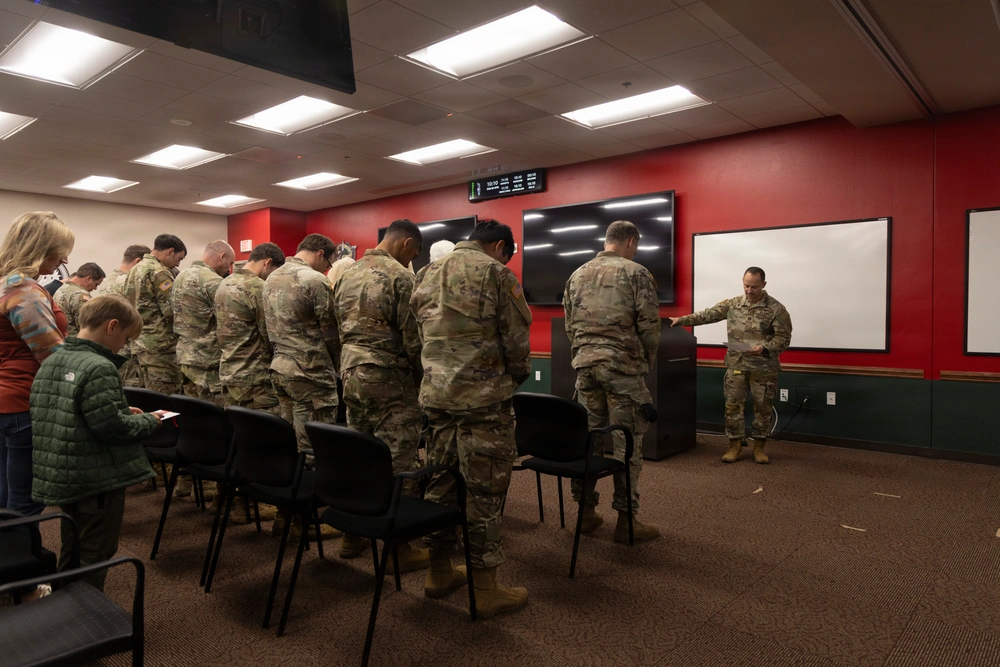 Soldiers in camouflage at a flag ceremony, led by an officer in uniform, in a room with a boy and woman watching.