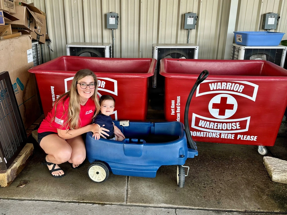 Volunteer with child by Red Cross bins at Fort Knox. "Warrior Warehouse Donations Here Please" text visible.