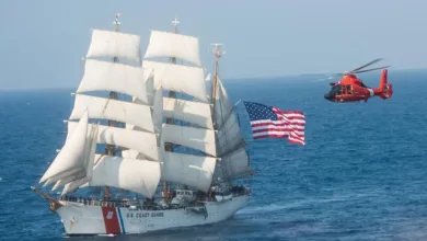 Coast Guard's Eagle sailing ship with American flag, accompanied by a red Coast Guard helicopter.