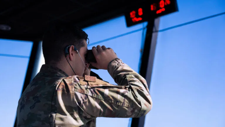 Air Force personnel in tower using binoculars, drone wingmen observation.