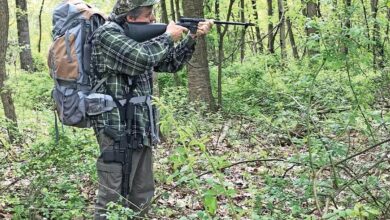 Man aiming Henry U.S. Survival Rifle in woods, wearing backpack.