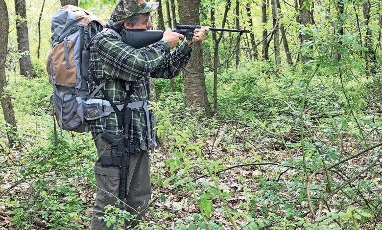 Man aiming Henry U.S. Survival Rifle in woods, wearing backpack.