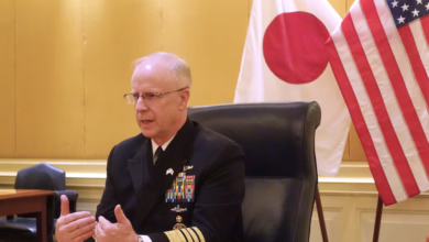 US admiral speaking with flags of Japan and the US behind him.