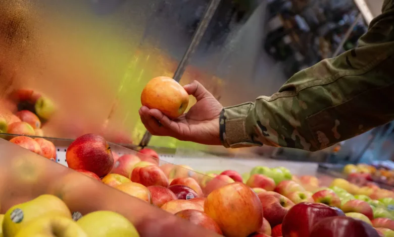 Hand in military uniform selects an apple from a display at a commissary.