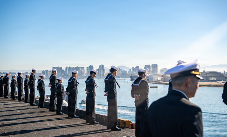 Navy sailors stand at attention aboard ship, San Diego skyline in background.