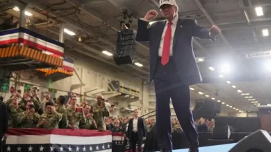 Donald Trump gesturing on stage at a rally with sailors. "USA" hat visible.