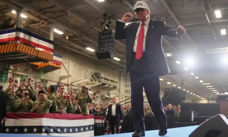Donald Trump gesturing on stage at a rally with sailors. "USA" hat visible.