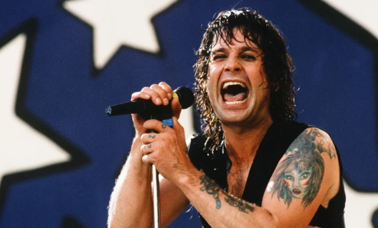 Ozzy Osbourne singing with a microphone at a concert, showing his arm tattoos.