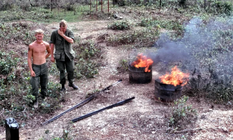 Two soldiers near burning barrels, possibly related to Vietnam burn pits, with one soldier appearing distressed.