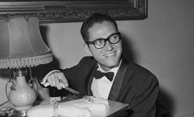Tom Lehrer cuts a cake, smiling in a black and white photo.