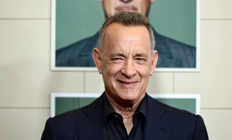 Tom Hanks smiling at an event, with a poster of himself in the background.