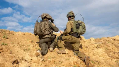 Two soldiers kneeling on a sandy hill wearing tactical gear and backpacks.