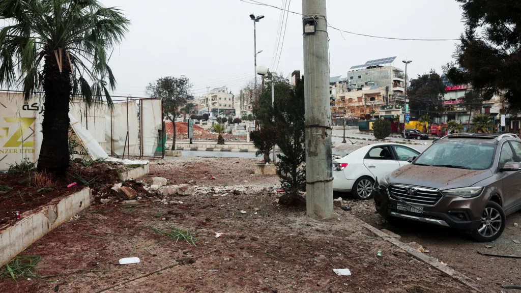 Street scene with damaged landscape, cars, and buildings under a cloudy sky.
