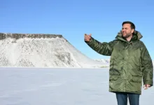 Man in parka gesturing in snowy landscape with mountain backdrop.
