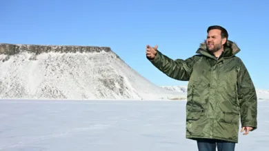 Man in parka gesturing in snowy landscape with mountain backdrop.
