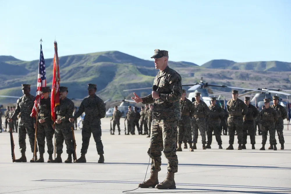 Marine officer speaking at HMLA-169 change of command ceremony, flags and helicopters visible.