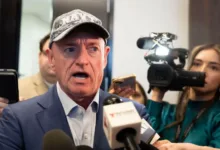 Mark Kelly speaks to press, wearing Navy hat and blue suit. Microphones visible.
