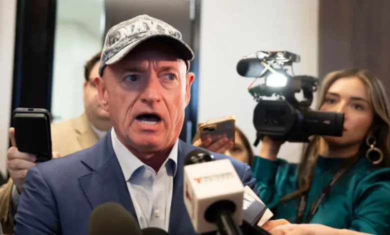 Mark Kelly speaks to press, wearing Navy hat and blue suit. Microphones visible.
