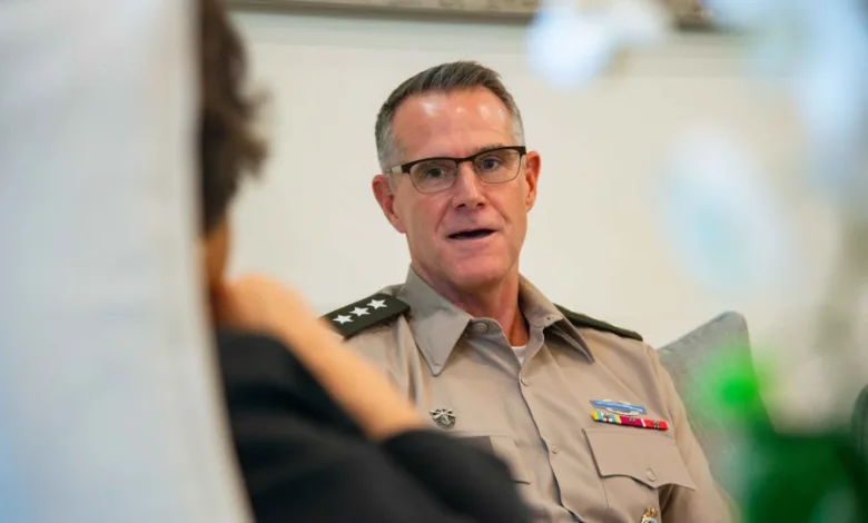 Man in military uniform with glasses speaking, seated indoors. Stars on shoulders, ribbons visible.
