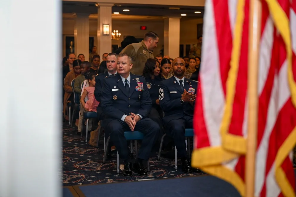 Military officers and audience at formal event, American flag foreground.