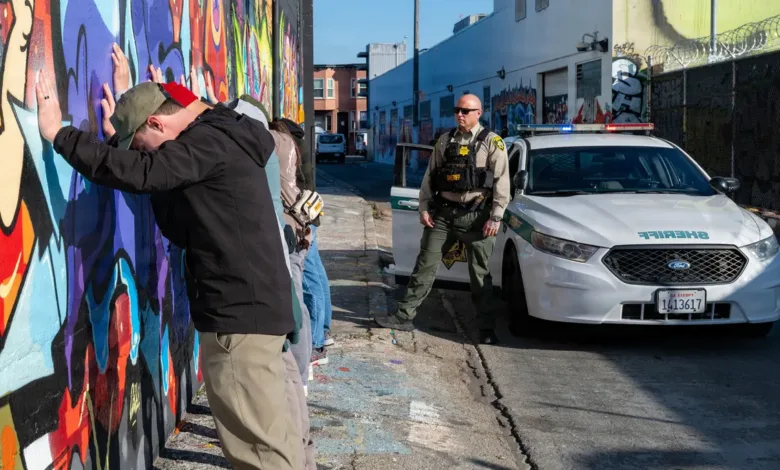 People against a mural wall being searched by a sheriff's deputy and patrol car during "Exercise Sourdough" in San Francisco.