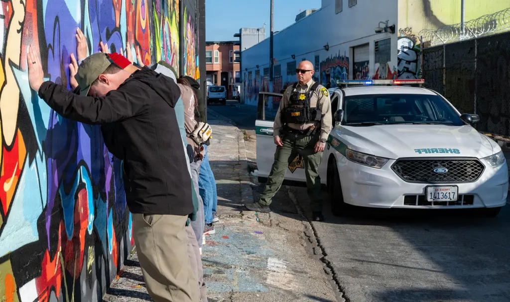 Man being searched against a mural by a sheriff near a patrol car in an urban environment.