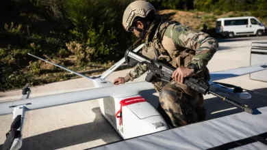 Soldier inspects drone with rifle, military gear, outdoor setting.