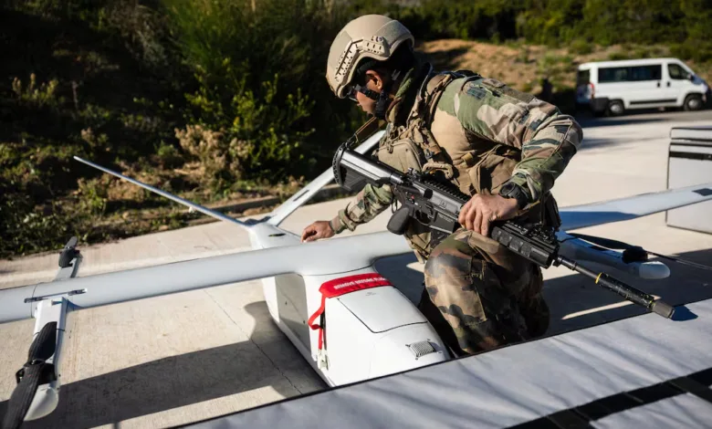 Soldier inspects drone with rifle, military gear, outdoor setting.