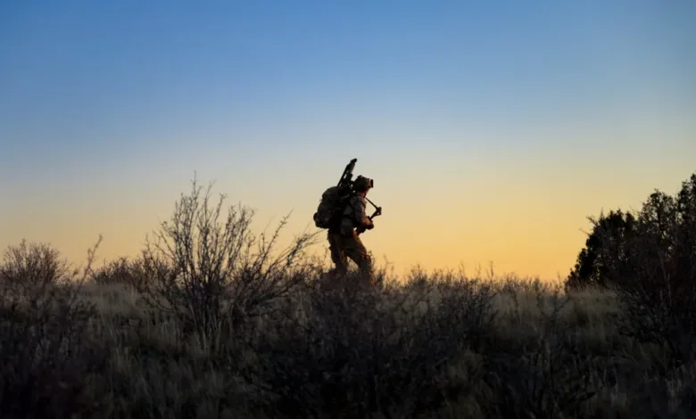 Soldier silhouetted against sunset, carrying gear through brush.