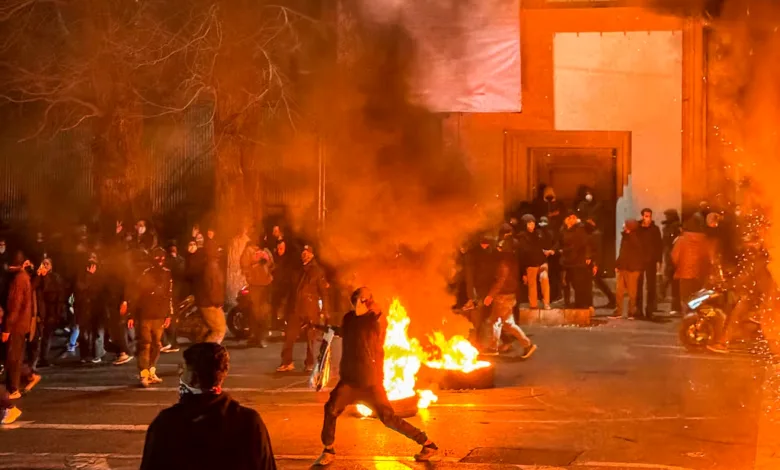 Protestors gather around a burning barricade in a street at night, creating a fiery scene with smoke and sparks.