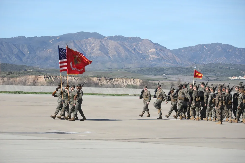 US Marines in formation march with flags, mountains in background.