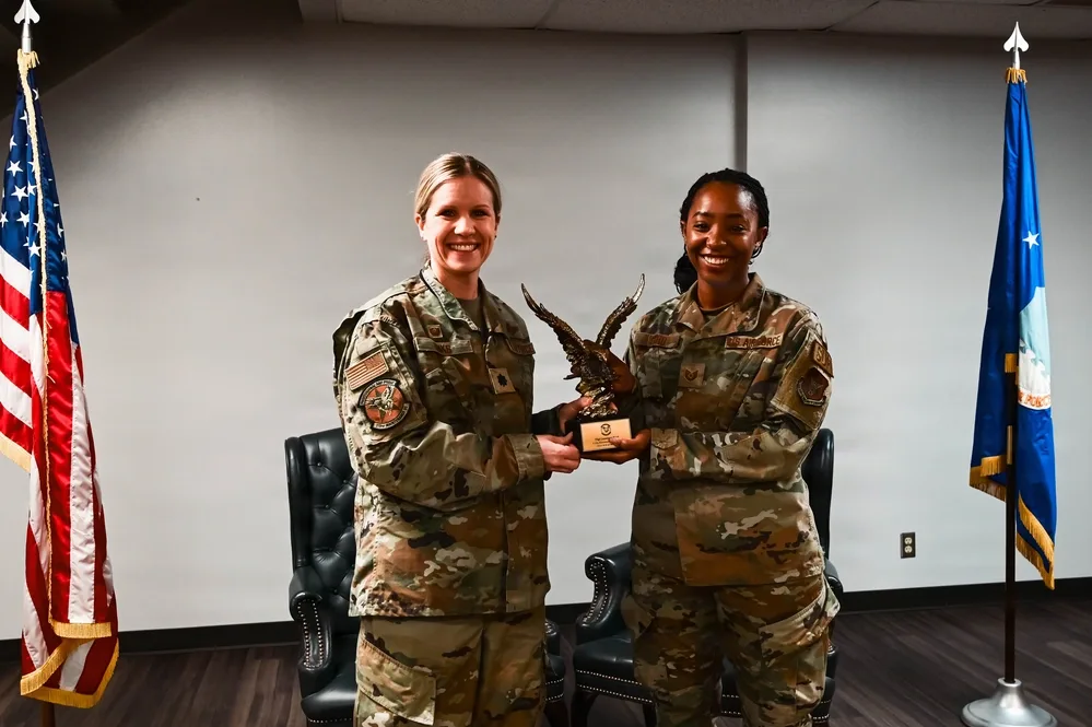 Two Air Force women holding award in office setting with US & Air Force flags.