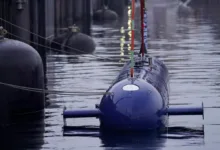 Blue submarine docked in water, front view. Buoys and dock in background.