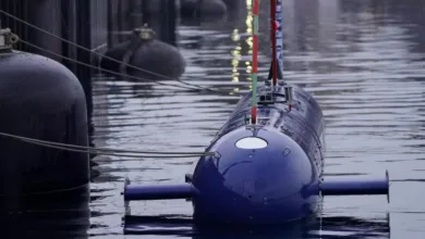 Blue submarine docked in water, front view. Buoys and dock in background.