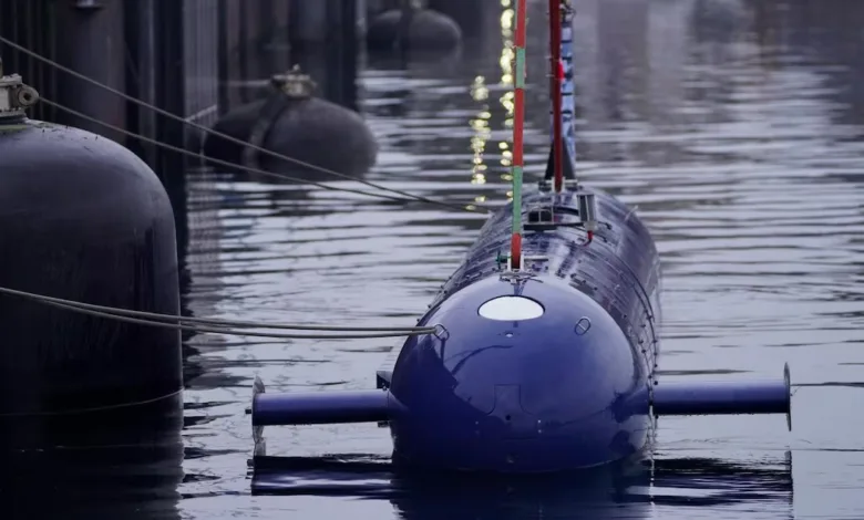 Blue submarine docked in water, front view. Buoys and dock in background.