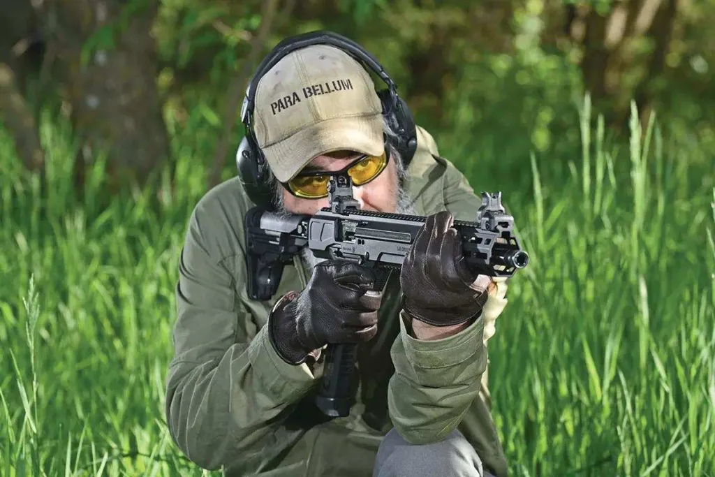 Man firing compact submachine gun with "Para Bellum" hat and ear protection.