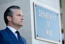 Man in suit by Department of War sign. Gray hair, serious expression.