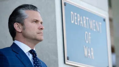 Man in suit by Department of War sign. Gray hair, serious expression.