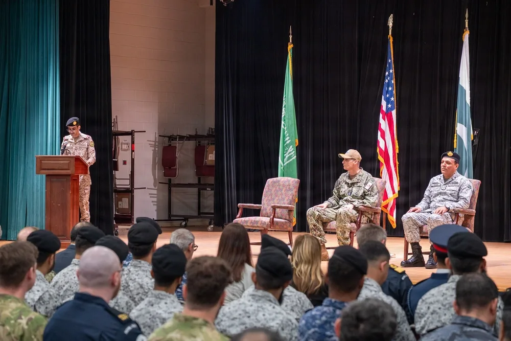 Military graduation ceremony with speaker, flags, and attendees in uniform.