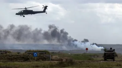 Military helicopter flying over smoky battlefield with armored vehicle and bunker in a field.
