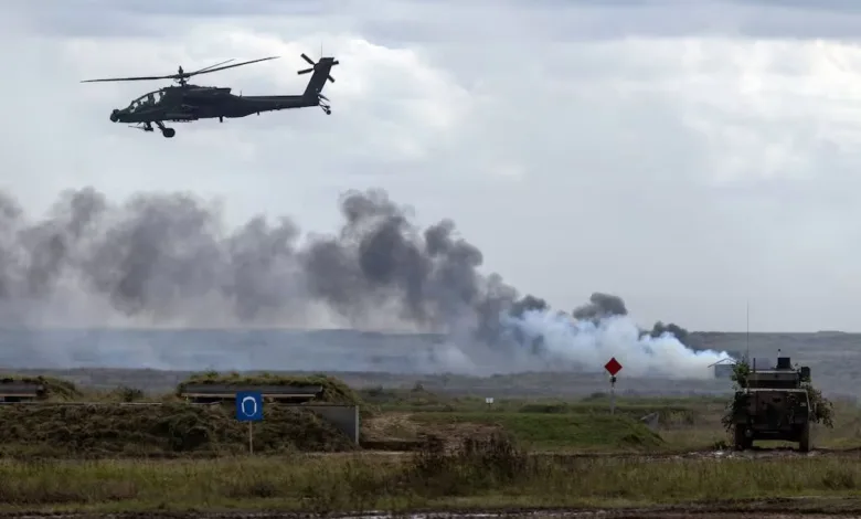 Military helicopter flying over smoky battlefield with armored vehicle and bunker in a field.