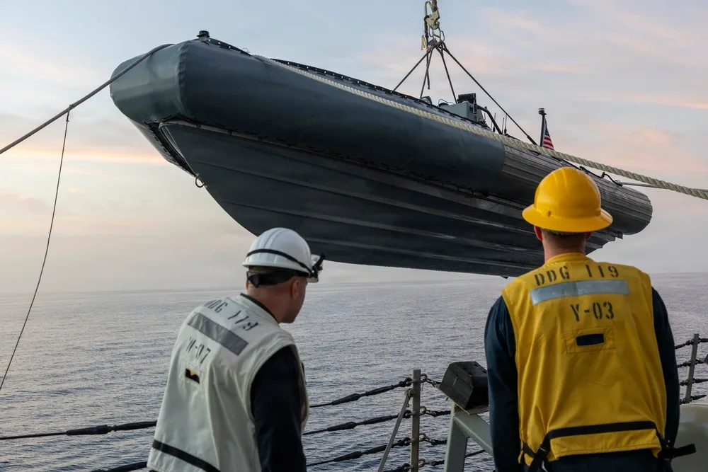 Rigid-hulled inflatable boat being lifted onto US Navy destroyer DDG 119, sailors watching.