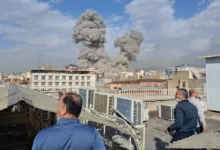 Smoke plumes rise over Tehran buildings as men watch from a rooftop.