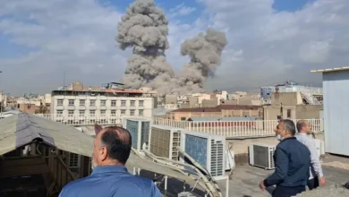 Smoke plumes rise over Tehran buildings as men watch from a rooftop.