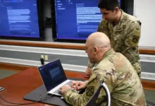 Two U.S. Army soldiers reviewing documents on laptop in a conference room.
