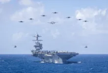 US Navy aircraft carrier with fighter jets and helicopters flying overhead in formation.