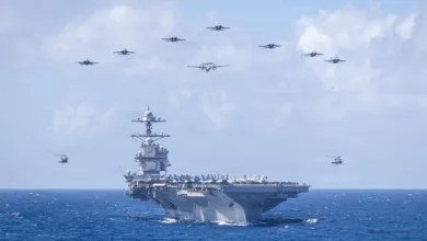 US Navy aircraft carrier with fighter jets and helicopters flying overhead in formation.