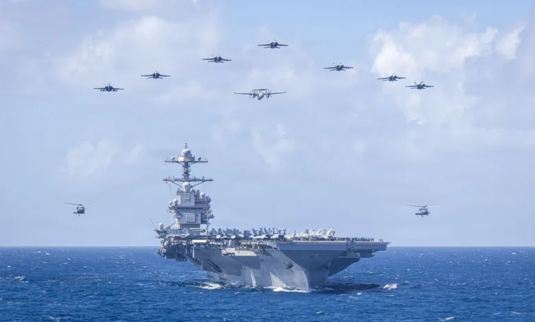 US Navy aircraft carrier with fighter jets and helicopters flying overhead in formation.