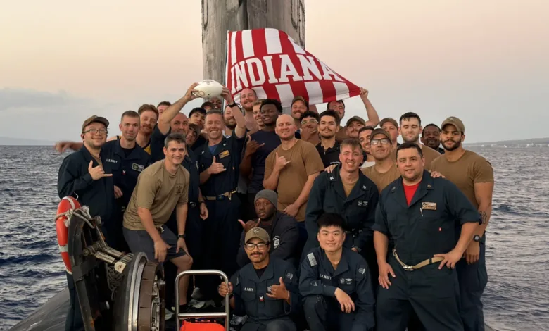 US Navy sailors on submarine with "Indiana" flag, smiling at the camera.